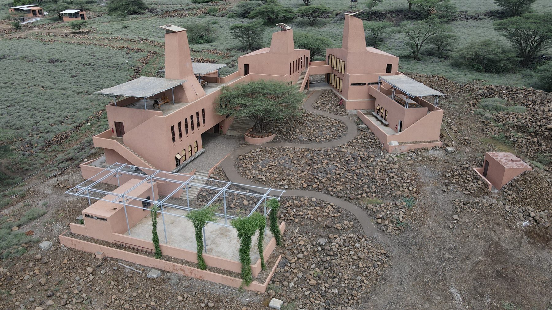 The Learning Lions campus from the air: three terracotta buildings with tall wind towers around an acacia tree, paths between the rocks, and the green plain of Lake Turkana's shore behind